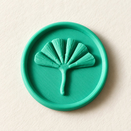 Detalle de un sello adhesivo redondo turquesa con hoja de ginkgo en relieve, acabado Minimal