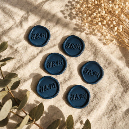 Seis sellos adhesivos azul marino con iniciales J&M en relieve, acabado rústico de borde irregular, sobre tela crema con eucalipto