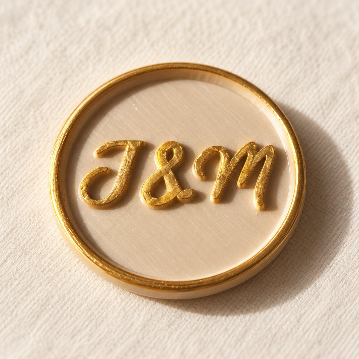 Detalle de un sello adhesivo arena con iniciales J&M doradas y borde dorado, acabado Signature