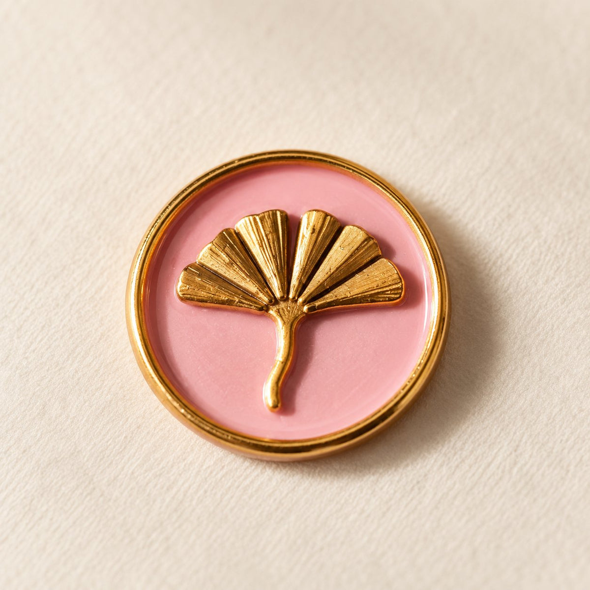 Detalle de un sello adhesivo de hoja de ginkgo dorada con borde dorado sobre base rosa, acabado Signature