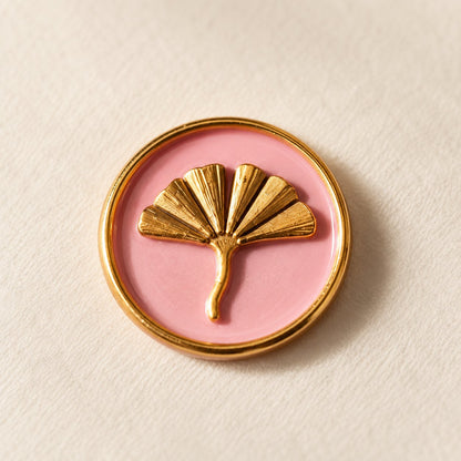 Detalle de un sello adhesivo de hoja de ginkgo dorada con borde dorado sobre base rosa, acabado Signature
