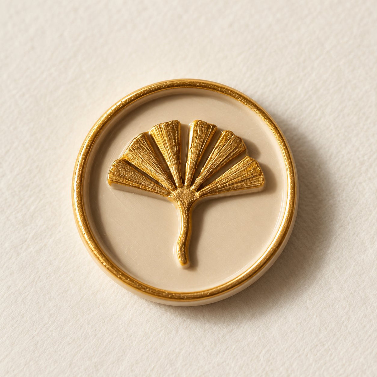 Detalle de un sello adhesivo de hoja de ginkgo dorada con borde dorado sobre base arena, acabado Signature