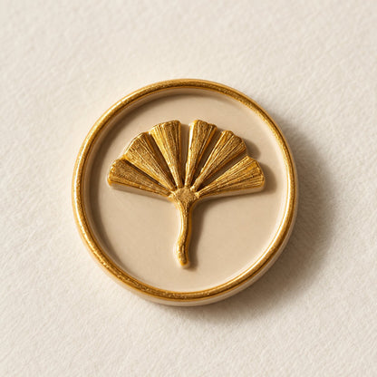 Detalle de un sello adhesivo de hoja de ginkgo dorada con borde dorado sobre base arena, acabado Signature