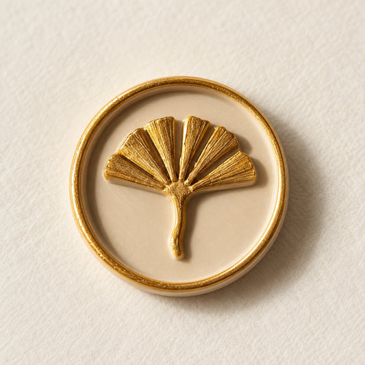 Detalle de un sello adhesivo de hoja de ginkgo dorada con borde dorado sobre base arena, acabado Signature