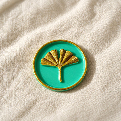 Detalle de un sello adhesivo de hoja de ginkgo dorada con borde dorado sobre base turquesa, acabado Signature