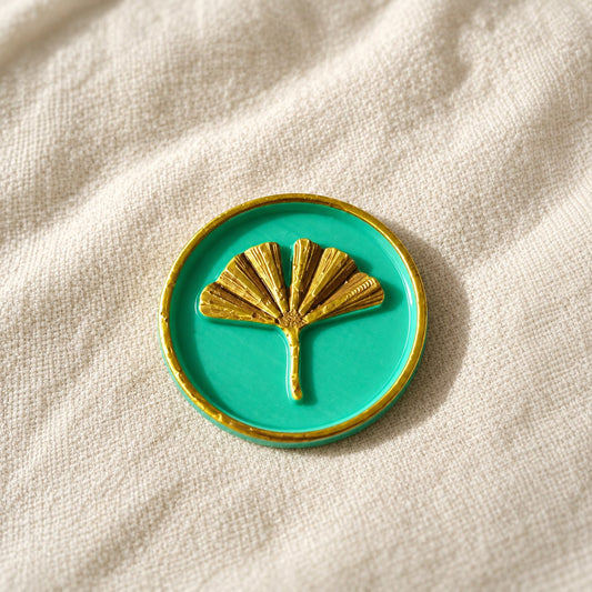Detalle de un sello adhesivo de hoja de ginkgo dorada con borde dorado sobre base turquesa, acabado Signature