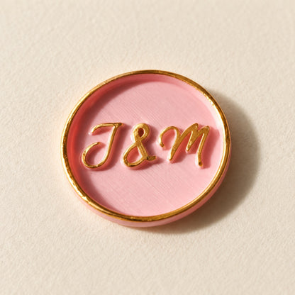 Detalle de un sello adhesivo rosa con iniciales y borde dorado pintado a mano