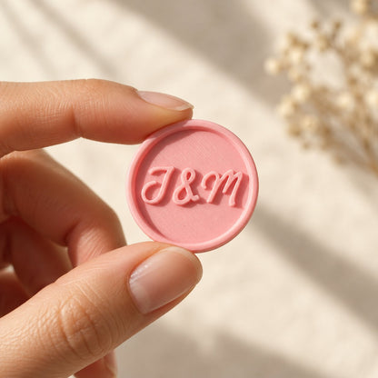 Mano sujetando un sello adhesivo redondo rosa con iniciales en relieve, acabado Minimal